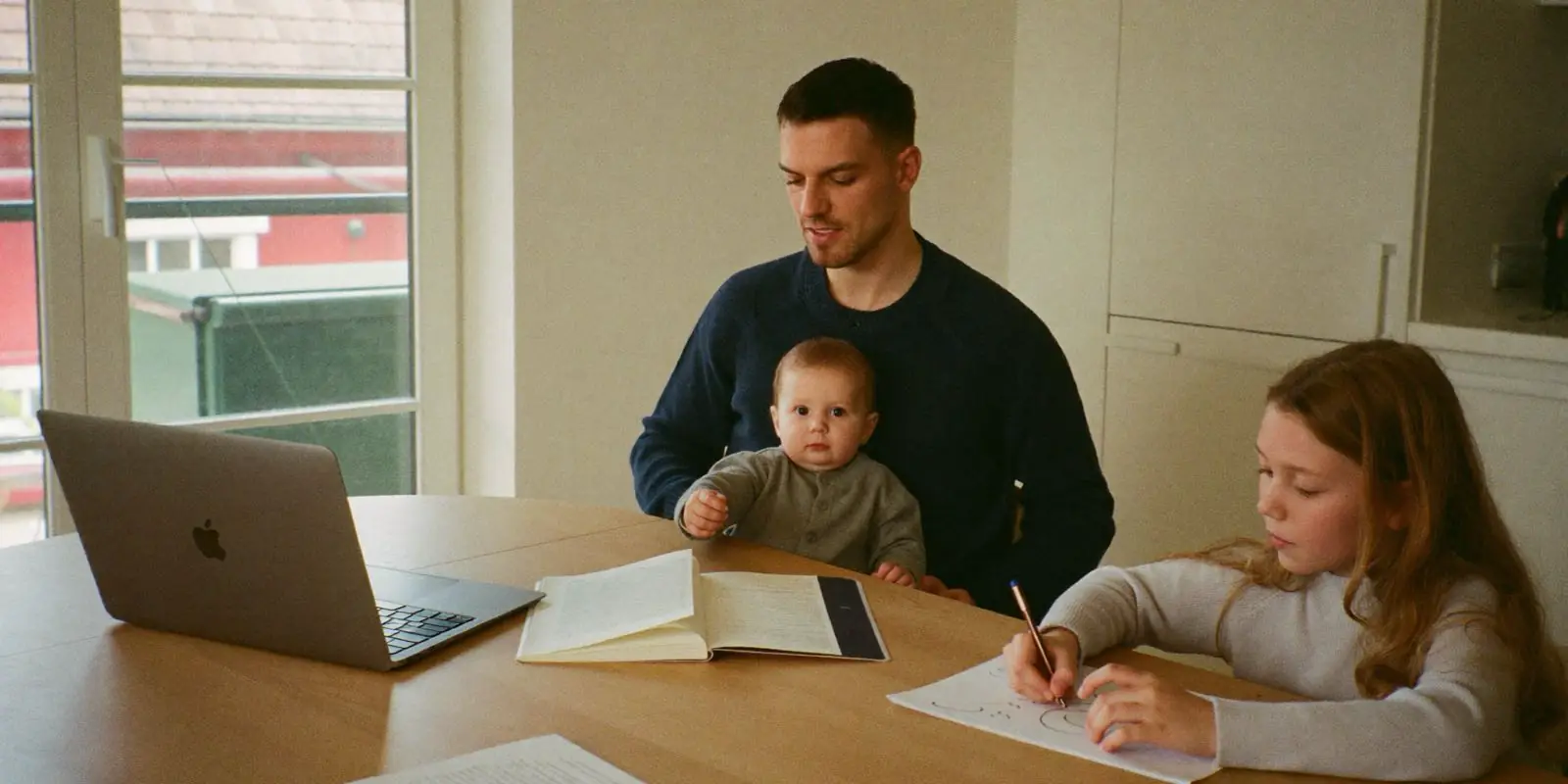Vater mit Baby auf dem Schoss und Tochter am Küchentisch mit Laptop und Unterlagen